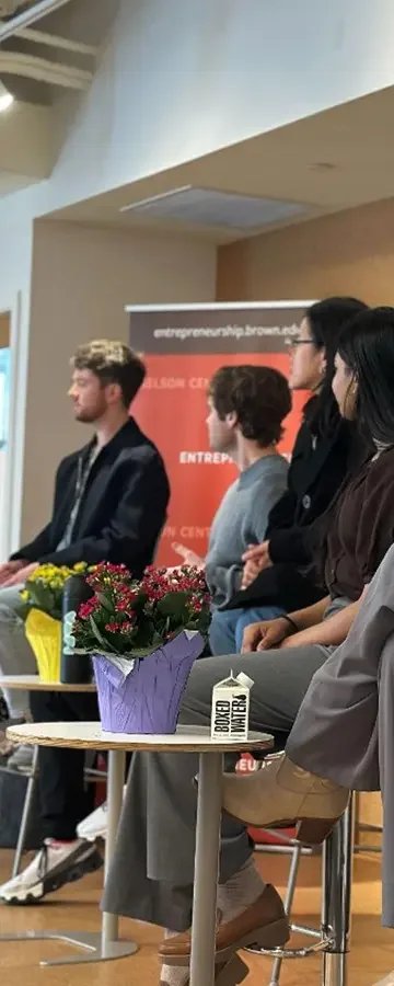 A panel of five people seated on stage during an event, with a red and black banner in the background that has the words 'entrepreneurship.brown.edu' and 'ENTREPRENEURSHIP'. In the foreground, there is a small table with a purple potted flower, a yellow item, and a white box labeled 'BOXED WATER'.