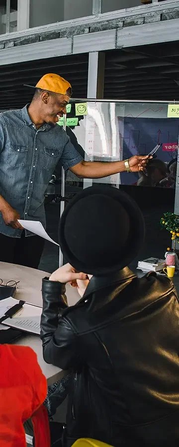 Two people in a meeting room with a man standing and pointing at a screen, while a woman in a black hat and leather jacket, seated at a table, looks on.