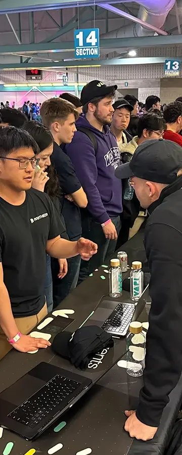 Group of people waiting in line at a booth, engaging with a man in black cap, with laptops and bottles on the table, inside a convention center.