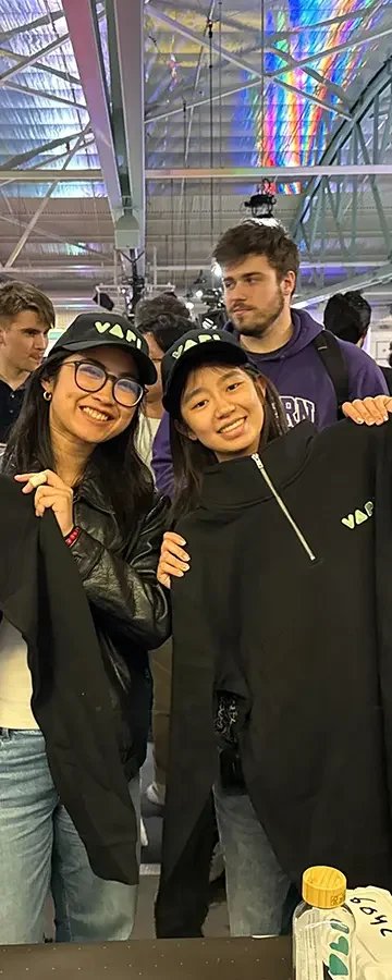 Group of young people smiling at a bean bag toss game, holding black jackets and wearing caps. The background shows an airport terminal with colorful lighting reflections on the ceiling.