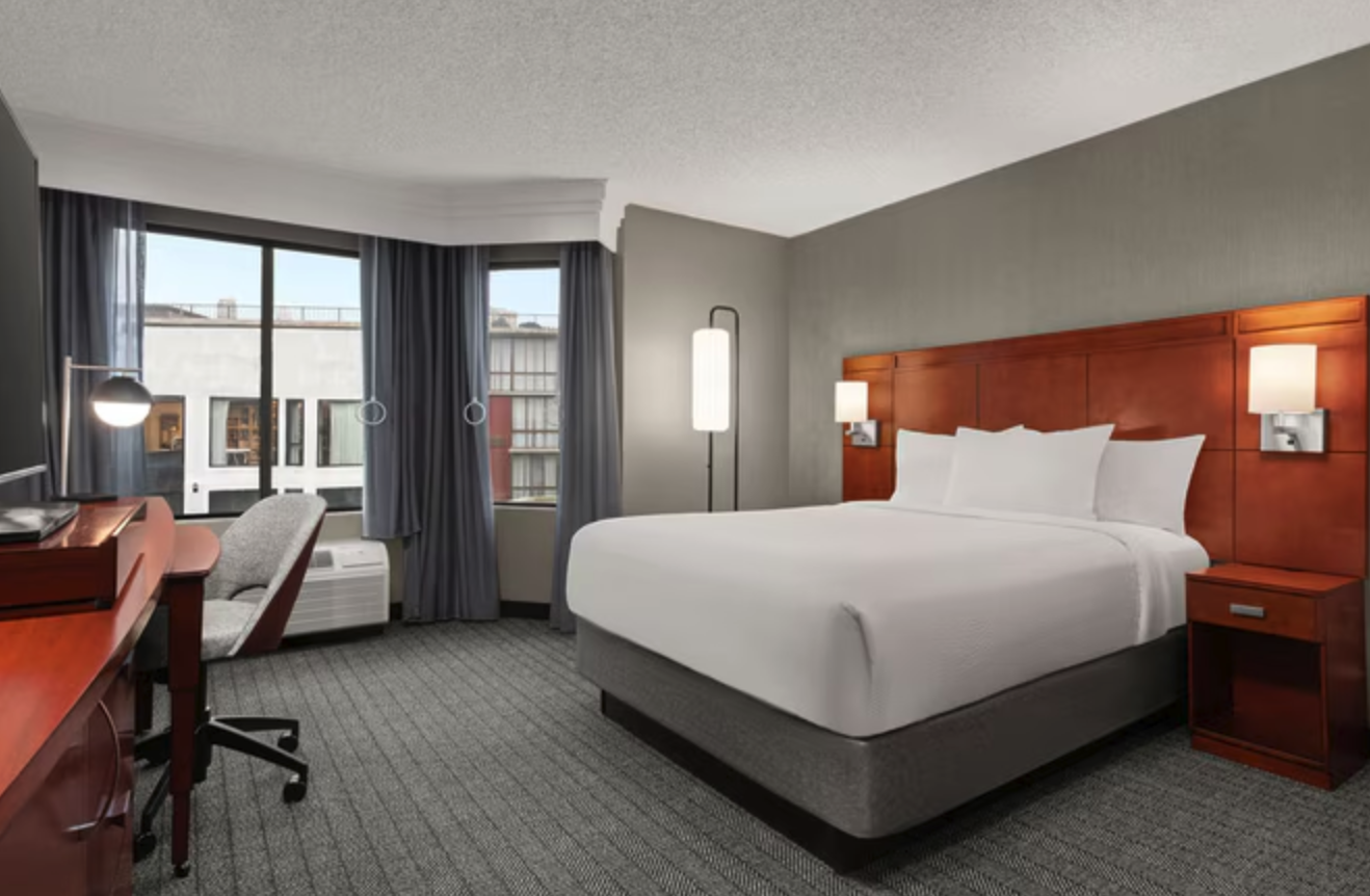A hotel room with a large bed, wooden headboard, and white bedding. There are two wall lamps, a floor lamp, a desk with a chair, windows with blue curtains, and city buildings outside.