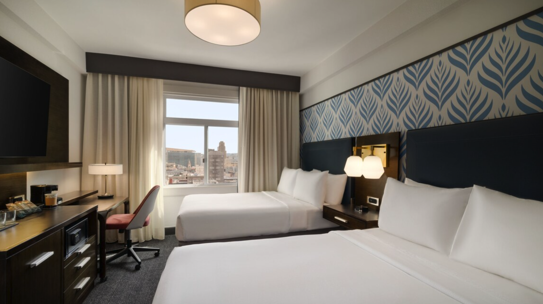 A hotel room with two white beds, a large window with city view, blue and beige patterned wallpaper, bedside lamps, a television, and a desk with a chair.