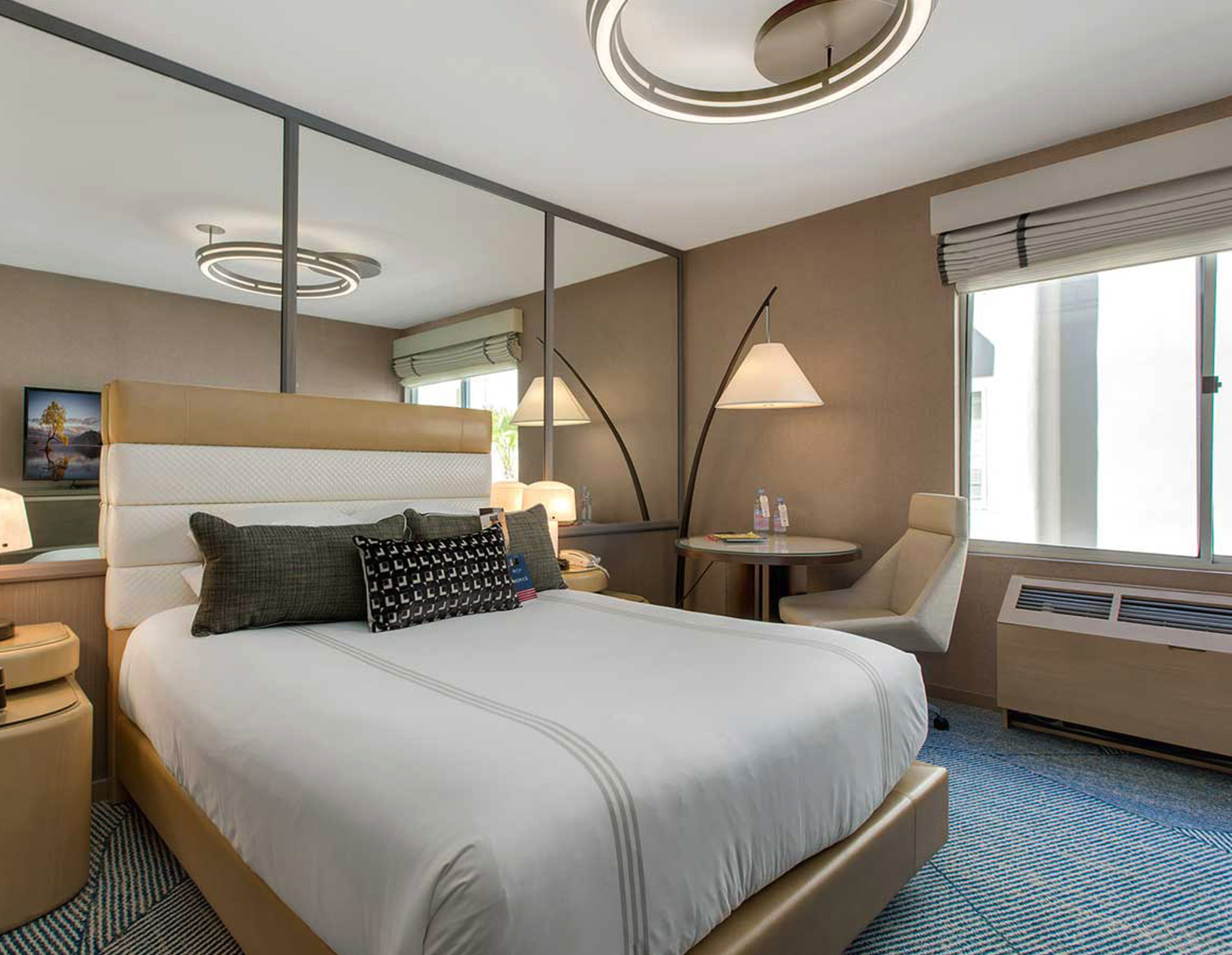 Hotel room with a large bed, pillows, bedside lamps, a mirror, a desk with two water bottles and a chair, and windows with blinds.