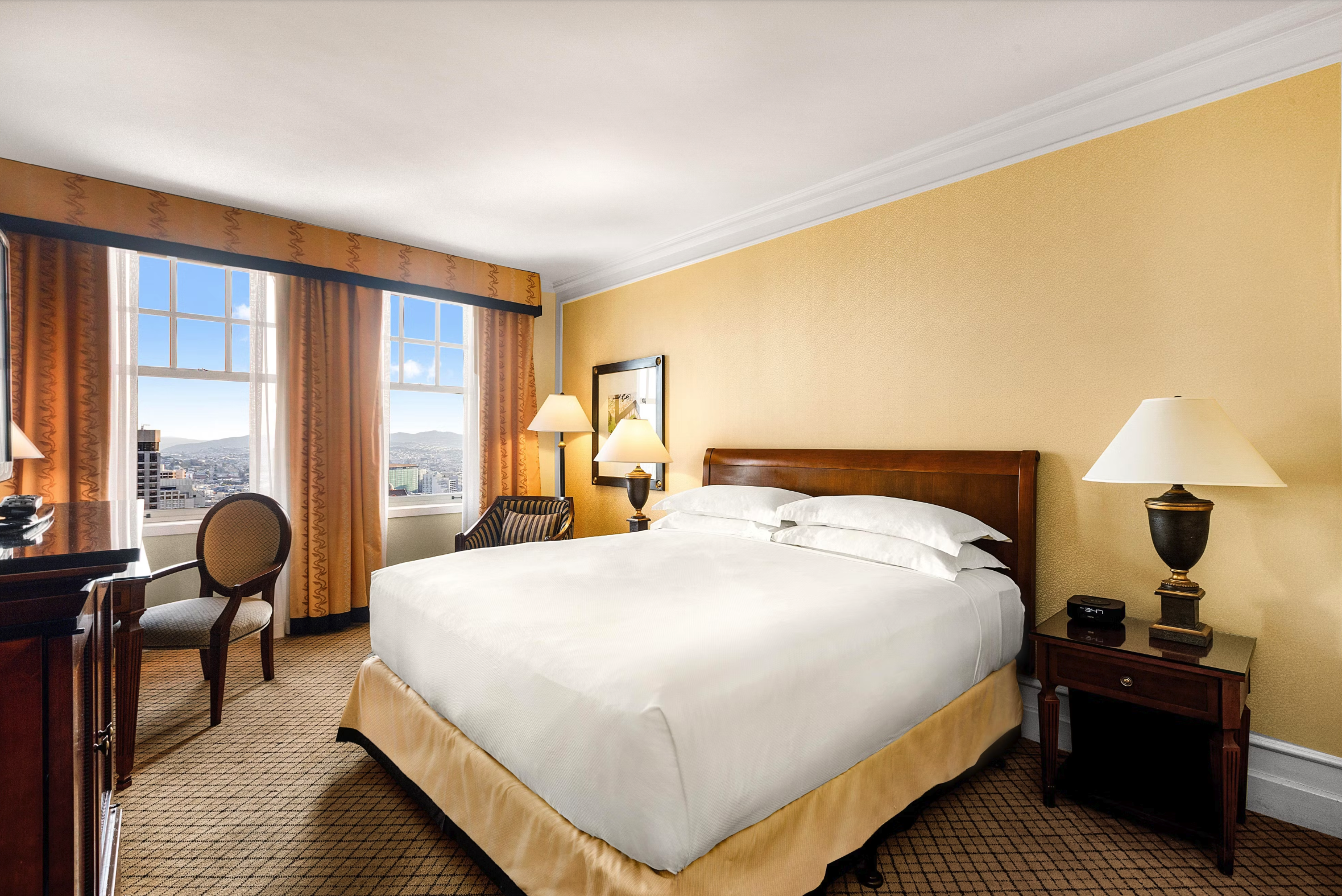 A hotel room with a large bed, nightstands with lamps, a framed picture on the yellow wall, a chair by the window, and city view outside.