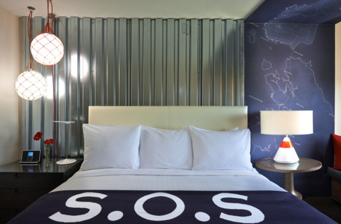 A hotel bedroom featuring a bed with white pillows and sheets, a dark blanket with 'S.O.S' written on it, two hanging spherical pendant lights, a bedside table with a phone and red flowers, and a wooden table with a modern lamp. There is a silver corrugated metal accent wall and a dark blue wall with a world map outline.